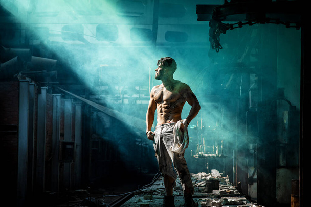 strong man against blue fog at factory exhausted stands, press and lateral abdominal muscles and pectoral muscles illuminated by light, place for text - Photo, Image