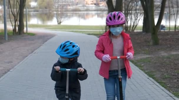 小さな男の子と女の子は公園の医療マスクのスクーターに乗っています。covid-19コロナウイルス感染からの保護の概念. - 映像、動画