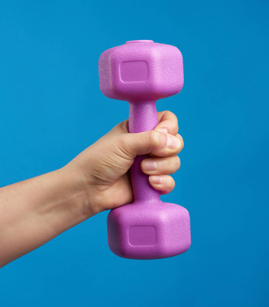 female hand holds a plastic pink dumbbell on a blue background, sport concept - Photo, Image