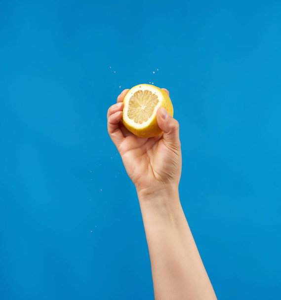female hand holds half a yellow lemon and squeezes it on a blue background, splashes fly to the sides - Photo, Image