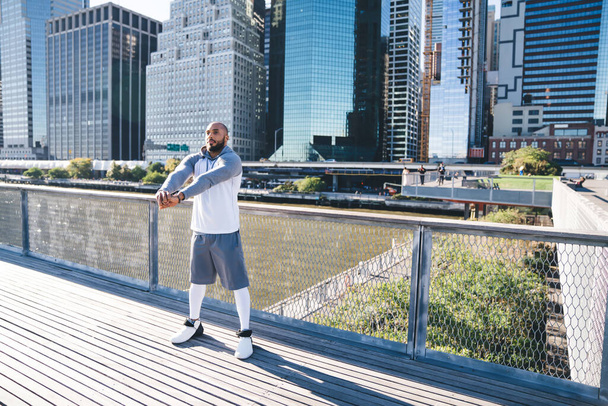 Bald bearded African American in sportswear stretching arms before running while standing on bridge during outdoor training with modern downtown in background - Photo, image