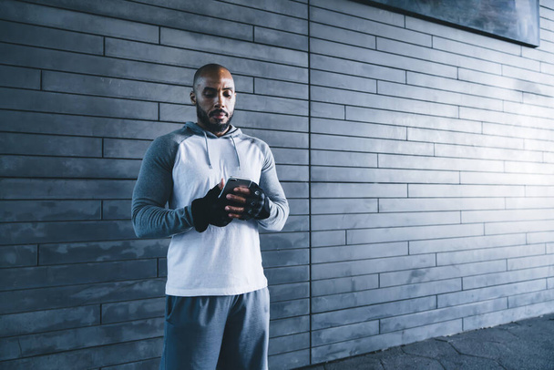 Athletic adult strong African American male in gloves surfing mobile while training in gym with gray wall on background in daylight - Foto, afbeelding