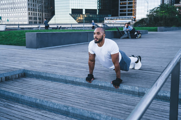 From above bearded focused ethnic male in sportswear and gloves having workout on promenade standing on hands and legs in plank pose looking away on background of buildings and people  - Foto, Bild