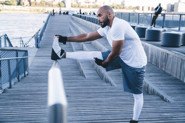 Bearded African American fit male in tracksuit and gloves practicing in morning time stretching legs on metal fence bridge looking away  - Фото, изображение