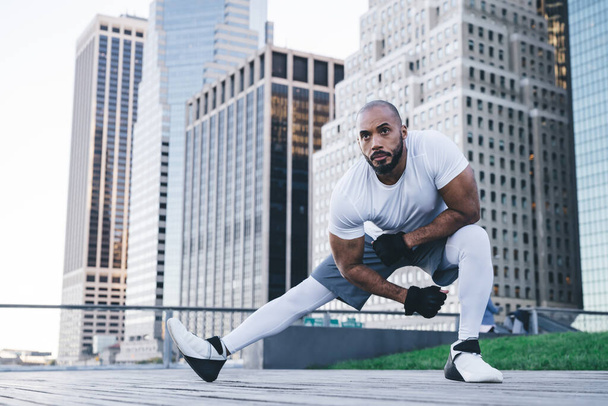 Bald athletic bearded African American guy in sportswear doing side lunge and stretching legs before training on streets of modern downtown - 写真・画像
