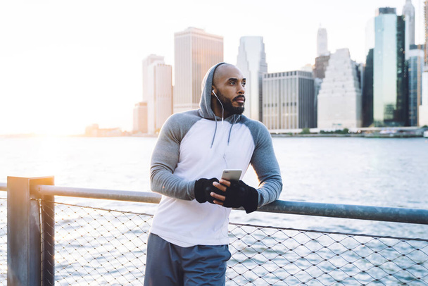 Bearded African American male in tracksuit and gloves holding phone listening to music leaning on metal fence on seafront in morning sunlight looking away - Fotografie, Obrázek