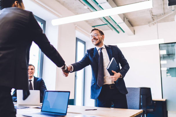 Happy male entrepreneur with notebook shaking hand of crop male business partner over table while being satisfied with successful negotiation meeting - Фото, изображение