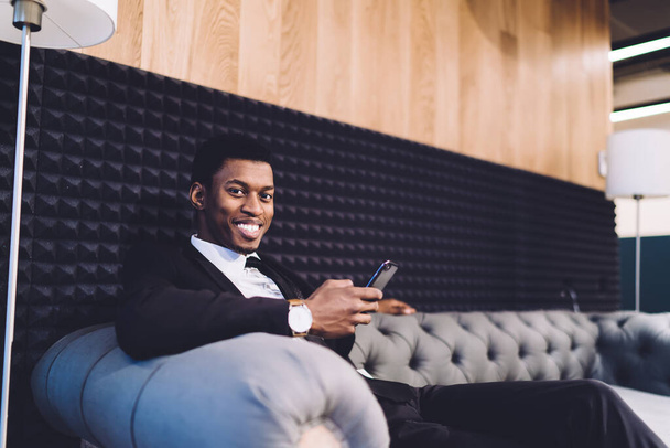 Cheerful African American male manager reading news on mobile phone while having rest and sitting on sofa in office lobby - Foto, immagini