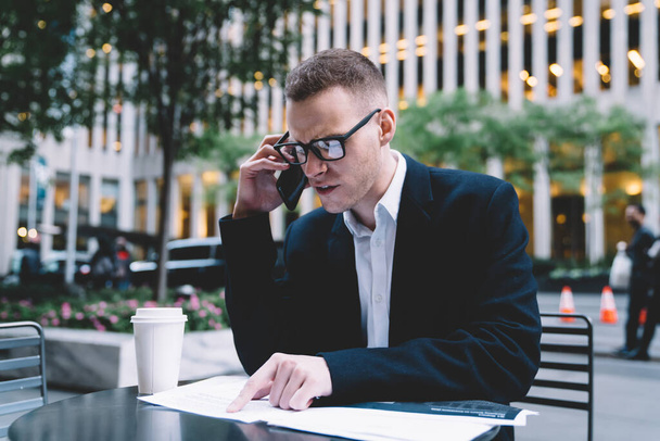 Focused businessman in black suit and glasses sitting at table in street cafe talking on phone while reading document looking dissatisfied with work - Foto, immagini