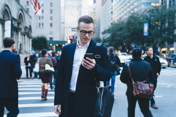Modern stylish short haired formal man checking mobile app standing on crosswalk in center of modern city with blurred background - Φωτογραφία, εικόνα
