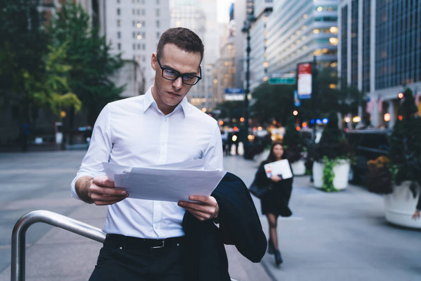 Handsome man in formal wear and trendy glasses attentively reading document while standing in middle of busy street in downtown and leaning on railing - Foto, Imagen