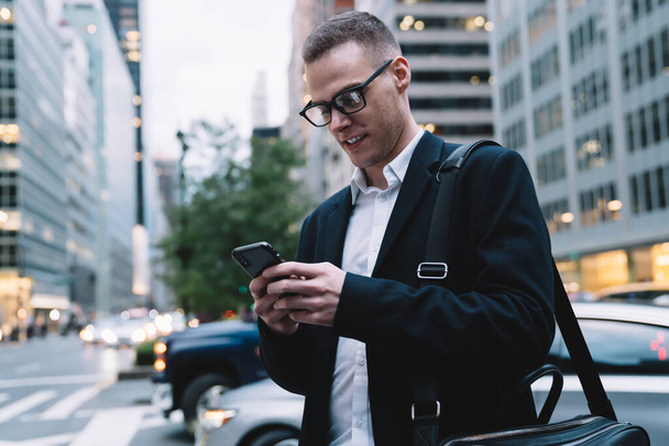 Smart and happy entrepreneur in formal suit and glasses walking around city and messaging in social media on blurred background - Fotografie, Obrázek