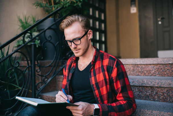 Focused  red haired male wearing checked red and black shirt making notes in notebook with pen sitting on granite stairs in city in summer - Foto, Imagem