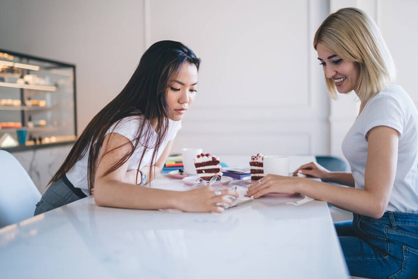 Multiracial smiling women in white t shirts looking at document sitting at table with pieces of cake cups of coffee notepads markers in cafeteria with counter in background - Foto, imagen