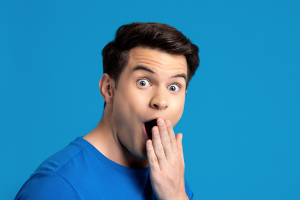 Retrato de modelo masculino caucasiano em gesto espantado com a mão cobrindo a boca em fundo estúdio isolado azul, seletivo focado
 - Foto, Imagem