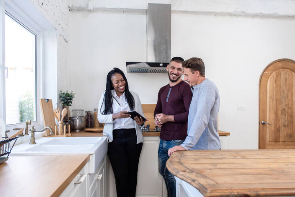 Africano americano feminino agente imobiliário na cozinha mostrando gay casal em torno de novo casa
 - Foto, Imagem