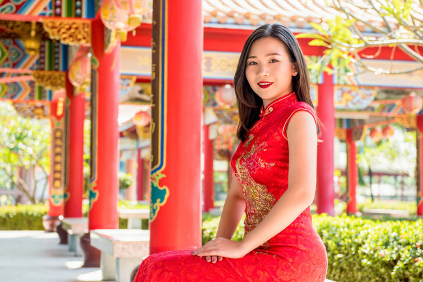 Smiling Asian woman in traditional red cheongsam qipao dress sitting with hands resting on lap in beautiful Chinese temple - Photo, Image