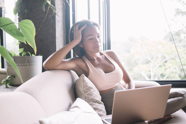 Dreamy youthful attractive Asian female in sporty clothes leaning on hand and looking away and holding laptop while sitting on sofa in comfortable flat - Fotó, kép