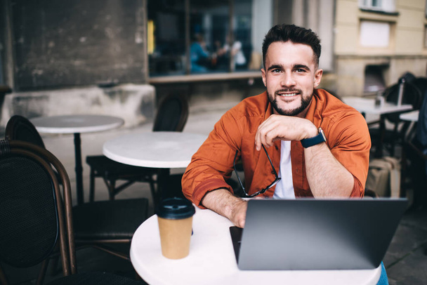 Estudante masculino otimista em roupas casuais com relógio inteligente trabalhando no laptop e sorrindo para a câmera enquanto sentado no café com café
 - Foto, Imagem