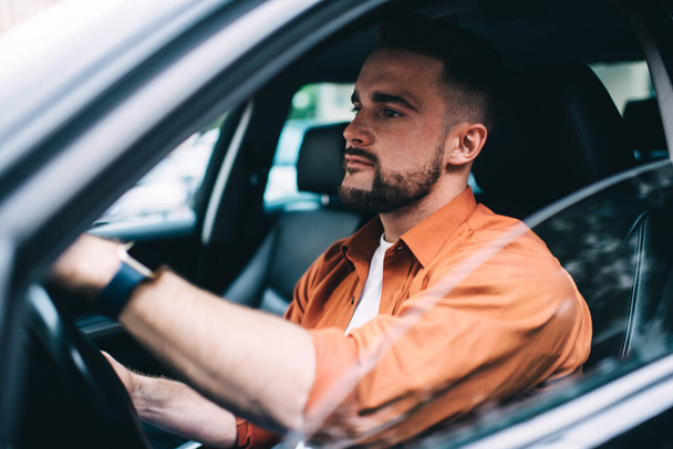 Side view of mindful bearded man in casual clothes sitting with hands on wheel in black car with rolled down windows looking away - Fotoğraf, Görsel