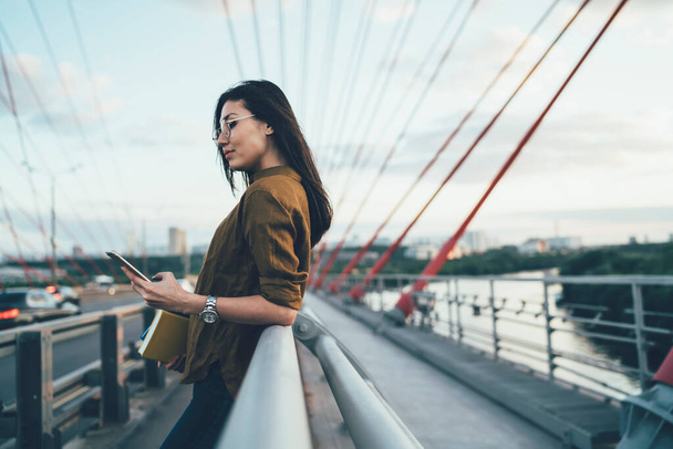 若いアジアの流行の服を着た女性は、都市の橋の上に休んでスマートフォンのソーシャルネットワークからのニュースを読んで、旅行中に共有出版物のための携帯電話を使用して女性旅行ブロガー - 写真・画像