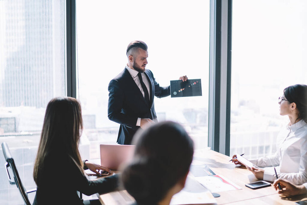 Bearded male executive in formal clothing with stylish haircut presenting chart to board of directors while standing near window in modern office building - Foto, imagen