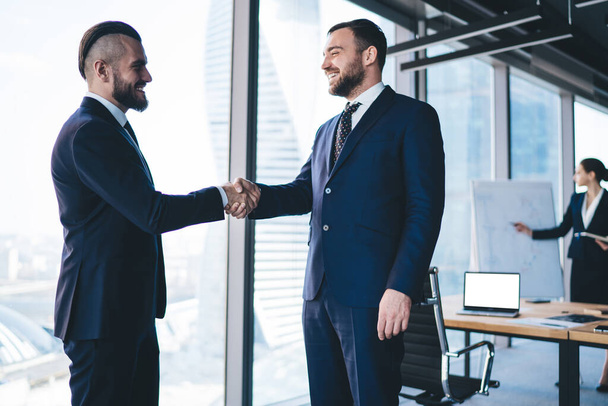Side view of pleased bearded businessman in formal clothes shaking hand of cheerful employee standing in office building near big windows - Fotoğraf, Görsel