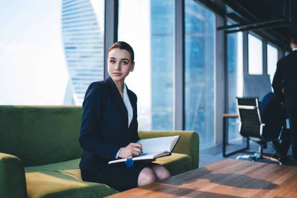 Young thoughtful female employee in formal suit writing notes in notebook while sitting on sofa in office building and looking at camera - Фото, изображение