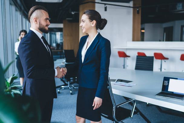 Side view of satisfied trendy entrepreneurs in business wear concluding deal in open space modern minimalist office and looking at each other on blurred background - 写真・画像