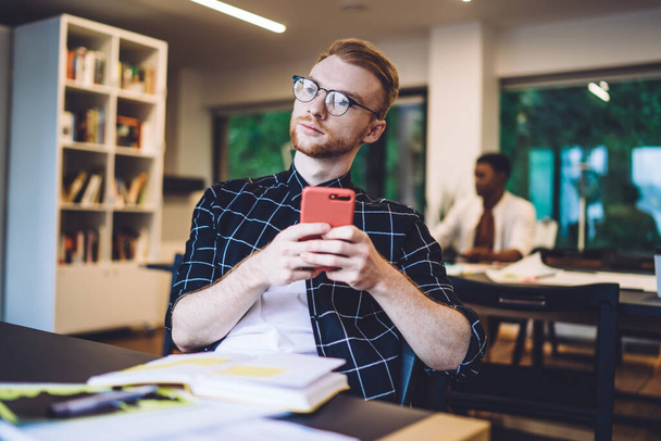 Thoughtful millennial blogger pondering on comments from personal website page during time for knowledge studying, young contemplative hipster guy in optical glasses holding modern mobile phone - Foto, Imagen