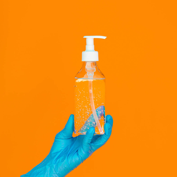 Uma mão em uma luva cirúrgica descartável azul segura uma garrafa de gel desinfetante contra um fundo laranja brilhante. Conceito de higiene e proteção à saúde durante a epidemia covid-19
 - Foto, Imagem