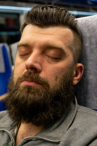 Mladý muž cestuje vlakem, který sedí u okna. Muž s plnovousem jede vlakem. Ten chlap spí na sedadle ve vlaku. Cestování. Vousatá bruneta. Muž v šedé bundě - Fotografie, Obrázek