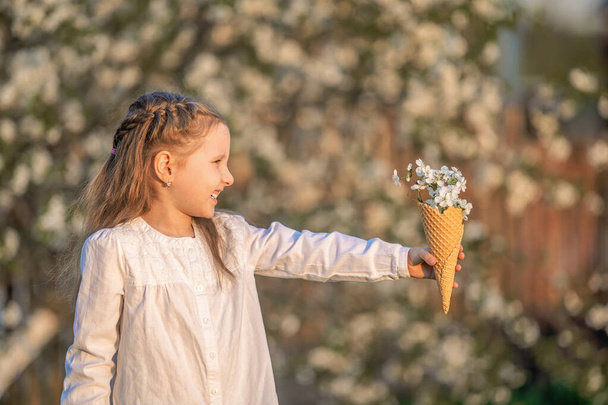 little laughing girl holds a waffle cone filled with cherry blossoms that stretches out, a side view, against a blurred natural background,at sunset. The concept of celebrating spring. - Foto, Imagen