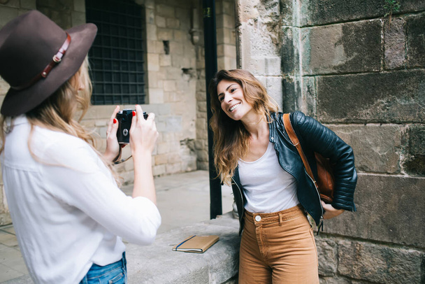 Anonymous blonde taking photo of smiled female student in casual stylish wear standing and smiling while posing for photo with stone wall on background - Φωτογραφία, εικόνα