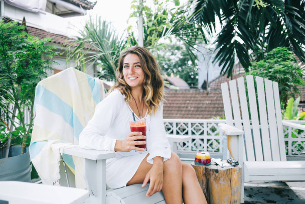 Optimistic satisfied young attractive female in white dress drinking red juice and relaxing on wood chair during holiday in tropical country - Foto, Imagem