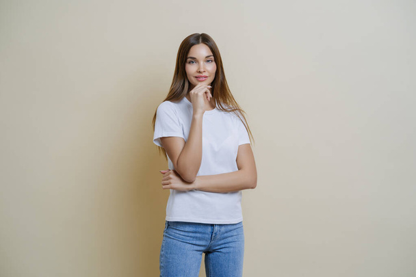 Photo of brunette young European woman holds chin, wears white t shirt and jeans, enjoys conversation with her mates, isolated over brown background. People, lifestyle, face expressions concept - Photo, Image