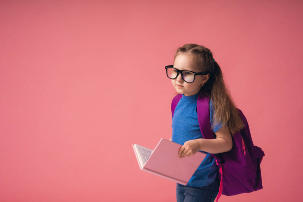 Cute, excited little girl in glasses with a school bag and holding a book goes to school, empty space isolated on a pink background, educational concept for school - Foto, Imagem