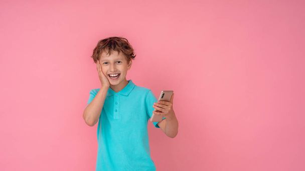 cheerful excited boy dressed in a Polo shirt, holding a smartphone on a pink background. Technology, children, the Internet. Close up. Copy space. caucasian - Foto, imagen