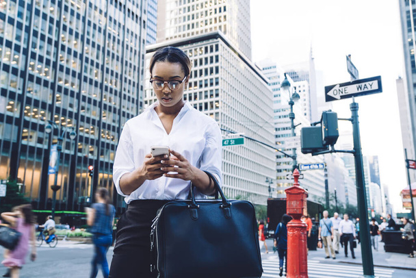 From below pensive serious African American businesswoman in glasses and formal clothes with black handbag focusing on screen and using smartphone while standing on street against blurred people and cityscape - Foto, Imagem