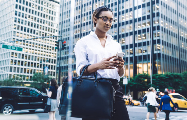 From below thoughtful African American confident woman in glasses and elegant clothes with black handbag focusing on screen and using smartphone while standing on street against blurred city life - Valokuva, kuva
