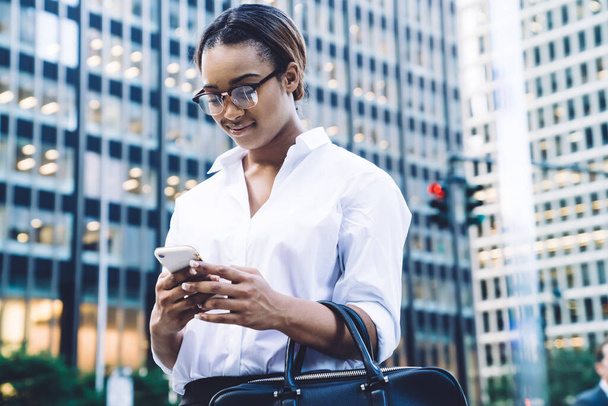 Cheerful successful young black female employee wearing glasses and white shirt chatting on smartphone while standing in street of downtown - Fotó, kép