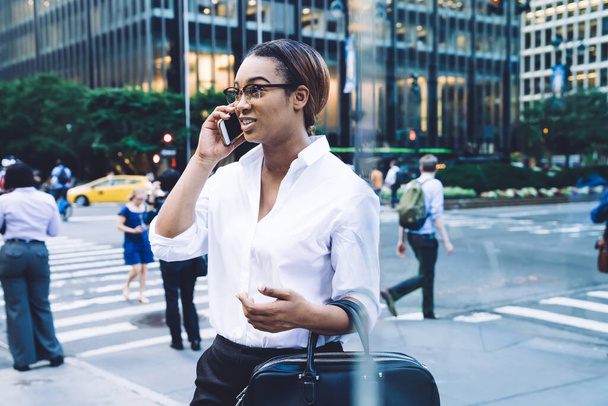 Modern confident African American woman in formal white shirt and eyeglasses talking on smartphone while standing on city street in downtown area - 写真・画像