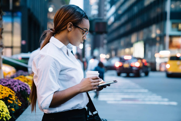 Side view of focused African American female with cup of beverage to go browsing modern tablet while standing near road on city street - Foto, immagini
