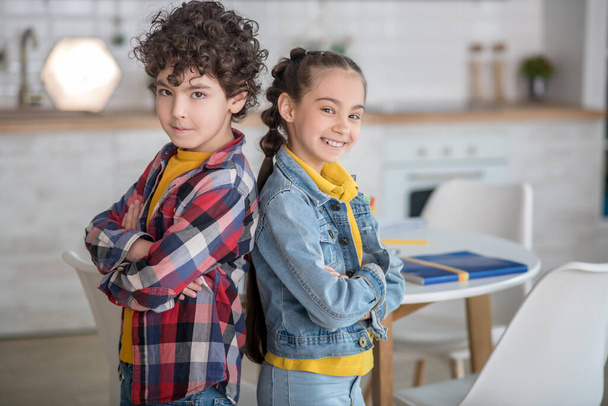 Dark-haired girl and curly boy smiling, standing back to back with folded arms - Foto, afbeelding