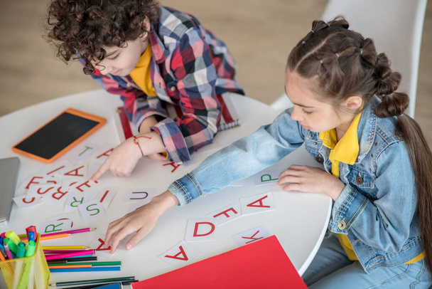 Curly boy and dark-haired girl sitting at round table, playing with alphabet letters - Photo, Image
