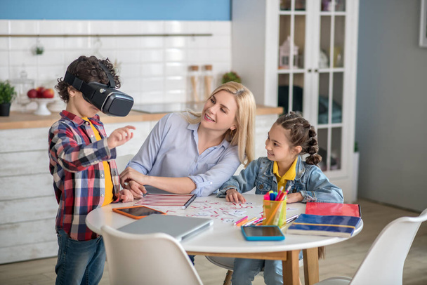 Curly boy and dark-haired girl sitting at round table with blonde female, boy wearing vr glasses, everybody smiling - Photo, Image