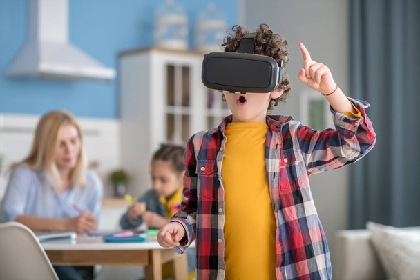Curly boy wearing vr glasses, looking excited, dark-haired girl and blonde woman sitting at round table, busy with tasks - Photo, image