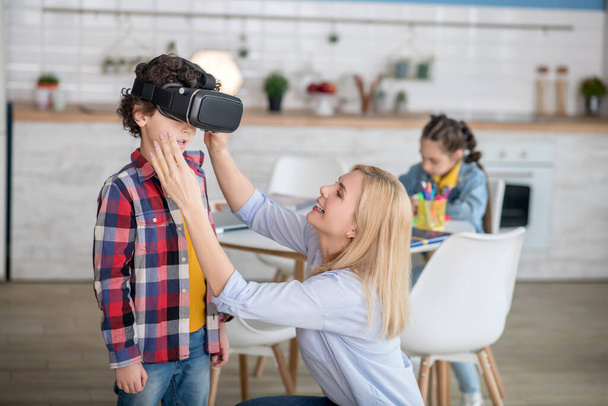 Blonde female putting on vr glasses on curly boy, dark-haired girl sitting at round table behind them, writing - Photo, image
