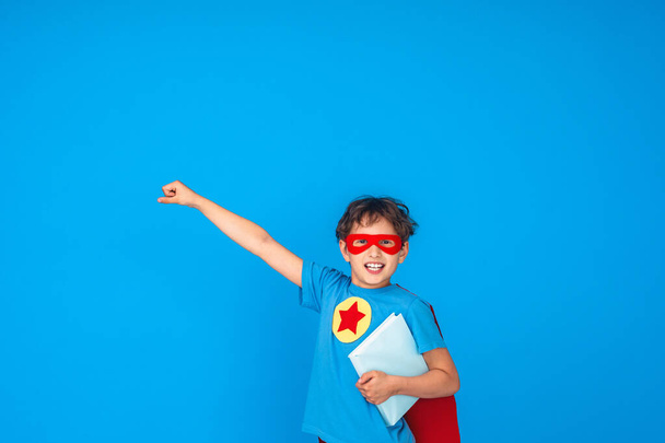 cheerful child dressed in superhero costume holds book and stretches out his hand. little boy laughs and enjoys learning at school. Posing on pink background in Studio Forward to knowledge. - Fotografie, Obrázek