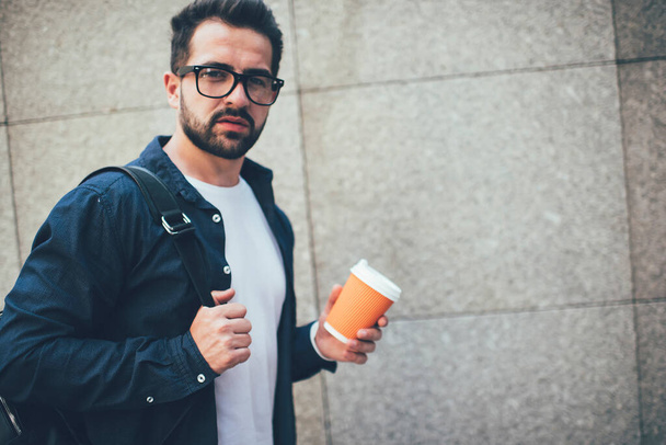 Half length portrait of serious caucasian hipster guy in casual wear and eyeglasses standing near wall outdoors, confident young 20s male student holding disposable cup with coffee takeaway  - Φωτογραφία, εικόνα
